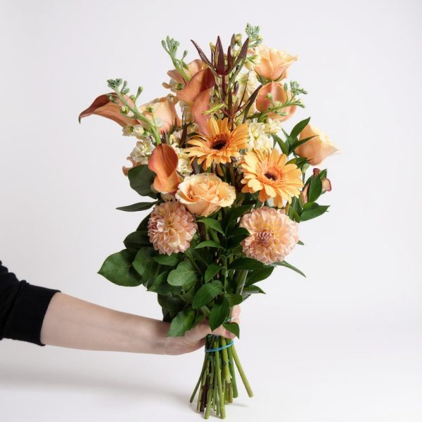 Golden Meadow bouquet held by hand with peach roses, dahlias, calla lilies, and gerberas in warm orange and cream tones