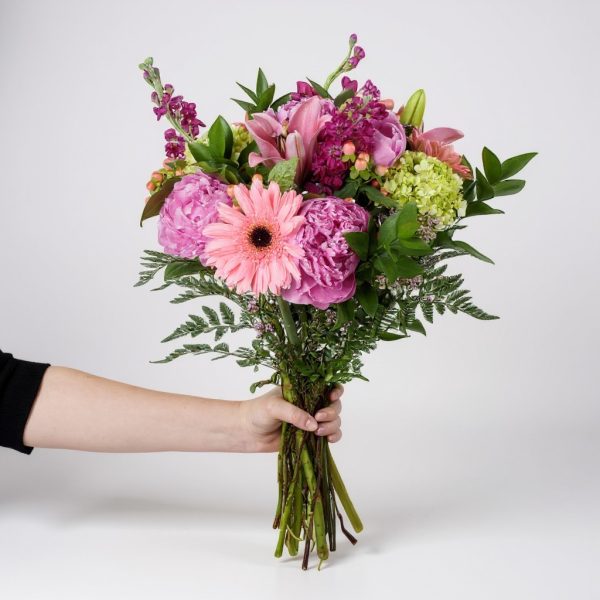 Hand holding bright pink bouquet with peonies, lilies, gerbera daisies, and green hydrangea