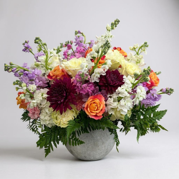 Colorful garden-style flower arrangement in a stone bowl featuring roses, dahlias, stock, and hydrangeas in warm and pastel tones