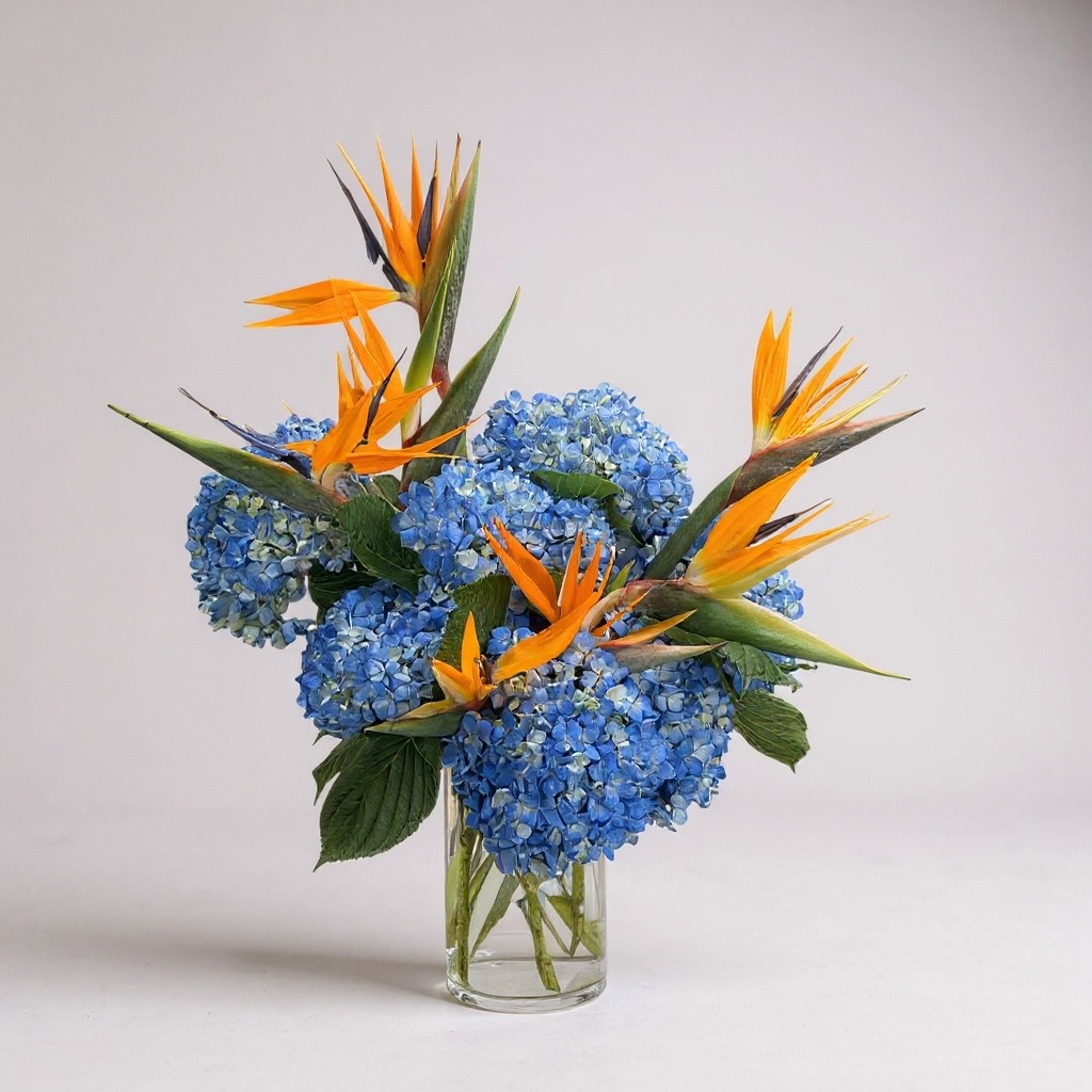 Modern vase arrangement with blue hydrangeas and orange Birds of Paradise in a clear glass vase