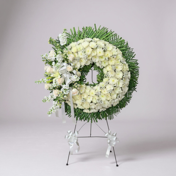 White funeral wreath with chrysanthemums, roses, lilies, greenery, and white ribbon bow on easel