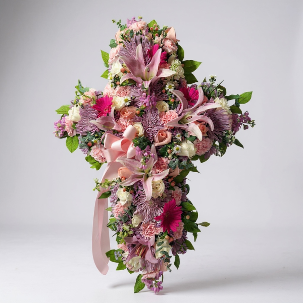Funeral cross spray with pink lilies, roses, gerbera daisies, pastel flowers, and pink ribbon bow on easel