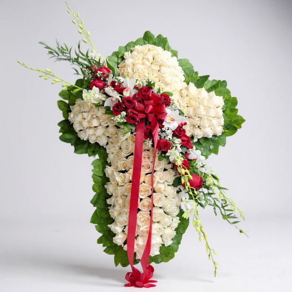 Funeral cross with white roses, red roses, orchids, greenery, and red ribbon bow on easel