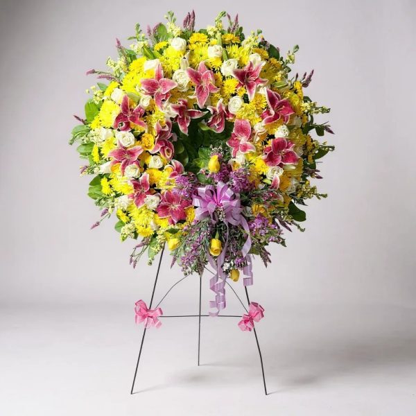 Funeral wreath with yellow chrysanthemums, white roses, pink lilies, purple flowers, and greenery with lavender ribbon on easel