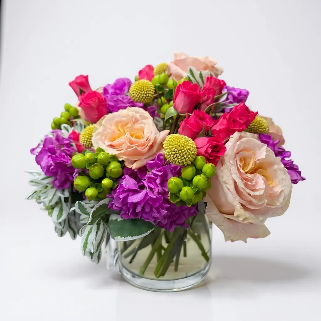 Colorful bouquet with peach roses, hot pink spray roses, purple stock, craspedia, and greenery in a glass vase