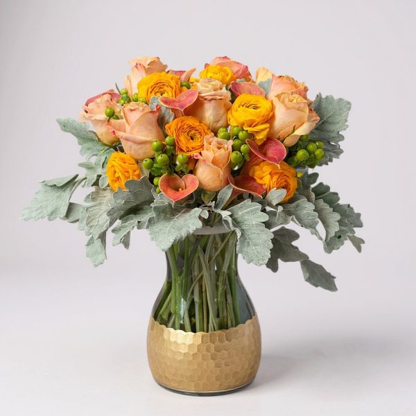 golden ember flower arrangement with peach roses, orange ranunculus, and dusty miller in gold honeycomb vase