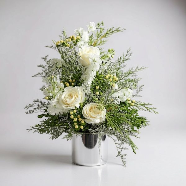 White Christmas flower arrangement with roses, frosted greenery, berries, and silver vase.