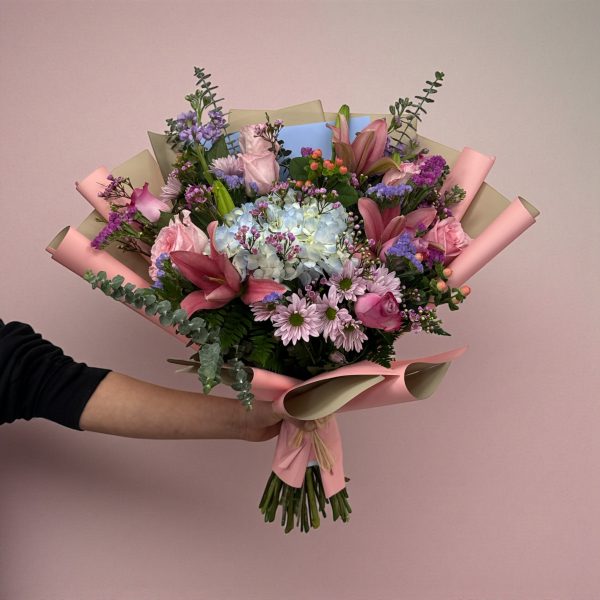 Hand-tied pastel flower bouquet with hydrangea, pink roses, lilies, daisies, and eucalyptus wrapped in pink and kraft paper.