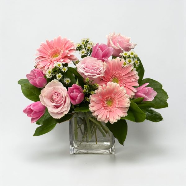 Mother’s Day arrangement with pink gerbera daisies, pink roses, tulips, and white accent flowers in a clear glass cube vase