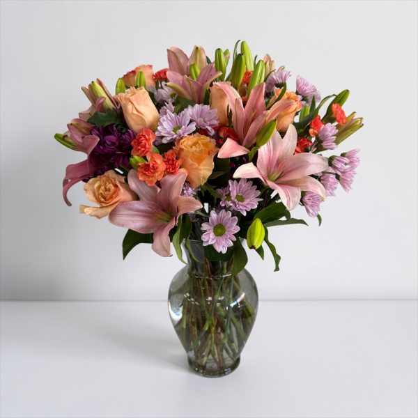 Pink lilies, peach roses, lavender daisies, and orange carnations in a clear glass vase
